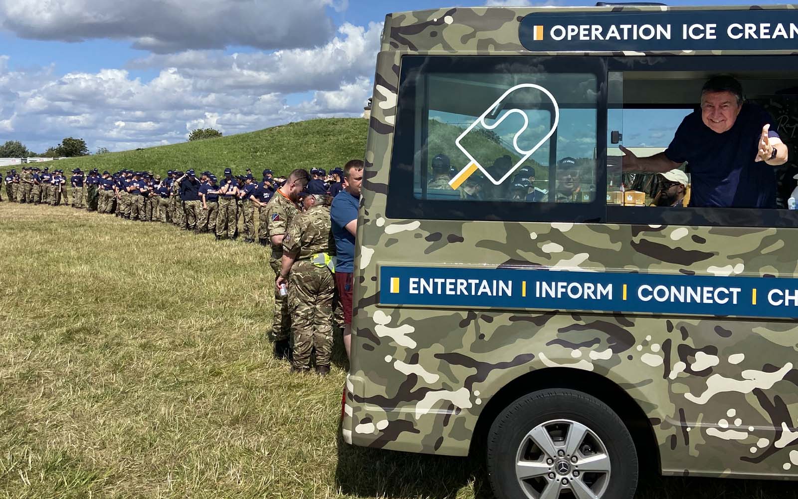 The BFBS Ice Cream van in a field with serving personnel queuing up to use it. 