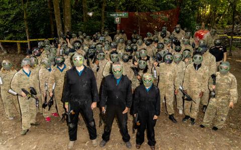 Group photo of those that took part in a paintballing event. Three stood at front in black overalls, several others in background with camouflage gear.