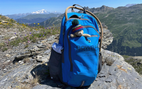 Close up of a back pack on a hill, scenic backdrop of mountains. Bag has a cute teddy bear sticking out of it.